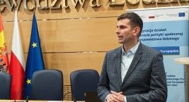 Mężczyzna w garniturze prezentuje się przed stołem z flagami.  