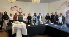 Ludzie stoją wokół stołu w sali, patrzą w stronę aparatu.