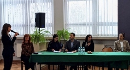 Grupa osób siedzi przy stole, a jedna osoba stoi obok z mikrofonem, prowadząc dyskusję