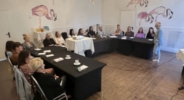 Wnętrze pokoju z kobietami siedzącymi przy stołach, które słuchają wypowiedzi osoby stojącej.