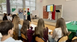 Osoby siedzą w rzędzie na krzesłach, zwrócone w stronę ekranu, uczestnicząc w wydarzeniu