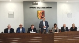 Grupa osób siedzących przy stole przed dużym znakiem