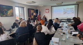 Uczestnicy siedzą przy stołach  i słuchają prelegentki prowadzącej prezentację